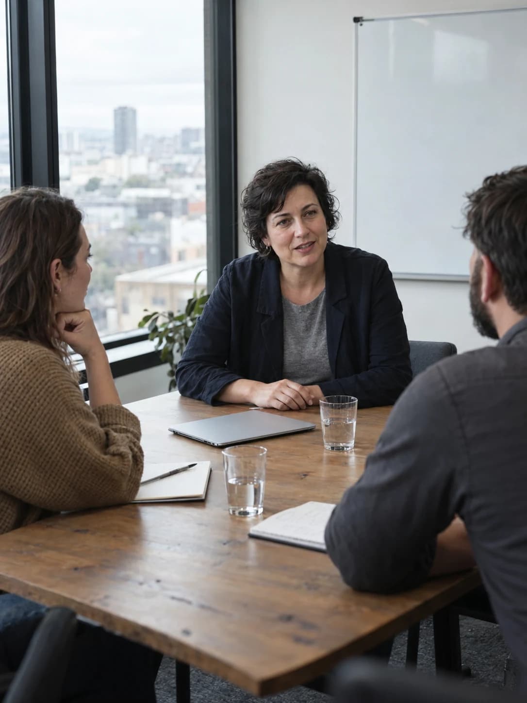 A trainer in a navy jacket leads a discussion with two colleagues at a wooden table in a window-lit meeting room.