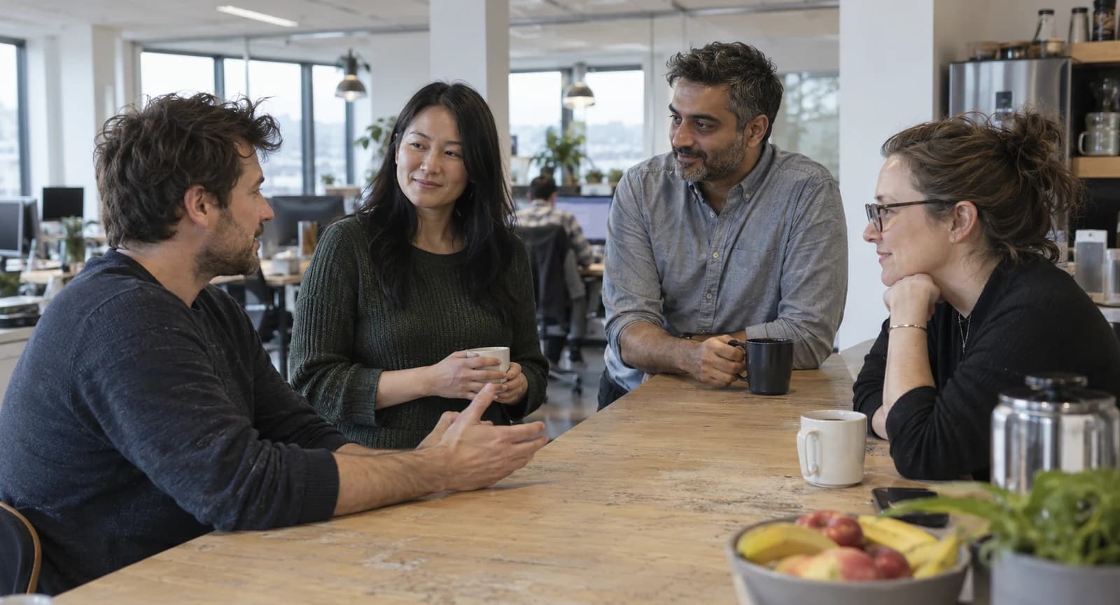 Four colleagues talk over coffee at a wooden counter in a bright office.